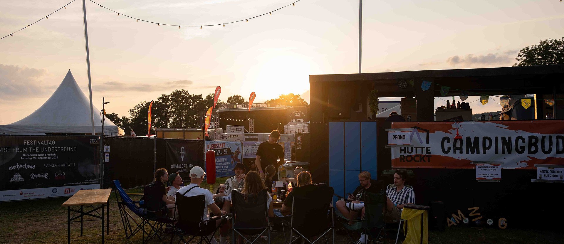 Besuchende des Festivals Hütte Rockt sitzen gemütlich in der Abendsonne zusammen. Im Hintergrund erkennt man die Hauptbühne
