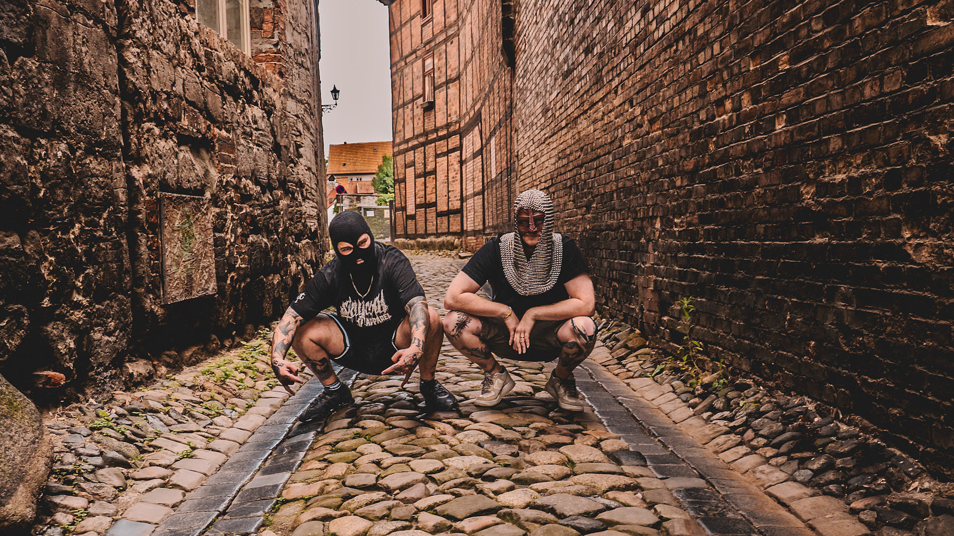 Die beiden Mitglieder der Band Mittel Alta hocken in einer Seitenstraße umgeben von Pflastersteinen und Steinmauern in mittelalterlichem Outfit