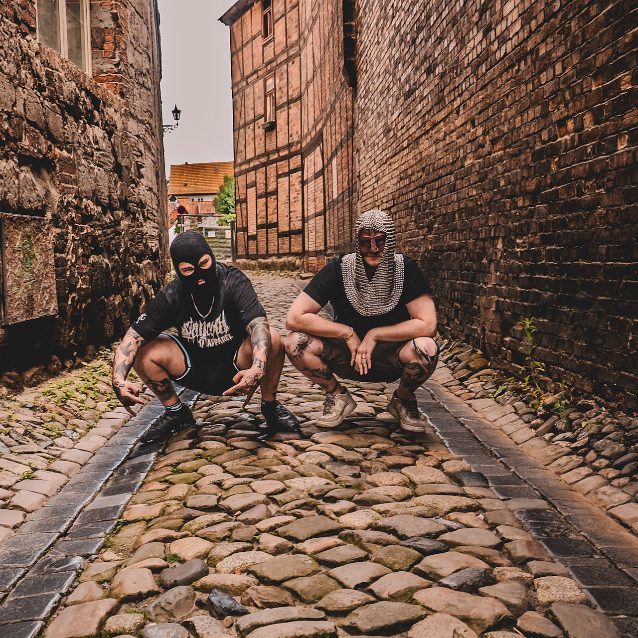 Die beiden Mitglieder der Band Mittel Alta hocken in einer Seitenstraße umgeben von Pflastersteinen und Steinmauern in mittelalterlichem Outfit