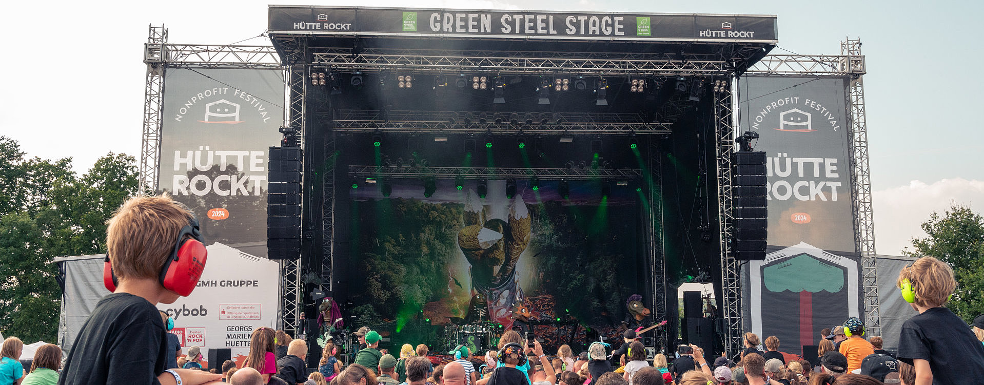 Auf der Hauptbühne des Hütte Rockt Festivals sehen wir die Band Heavysaurus. Davor steht eine große Menschenmenge und ein kleines Kind sitzt links auf den Schultern eines Erwachsenen