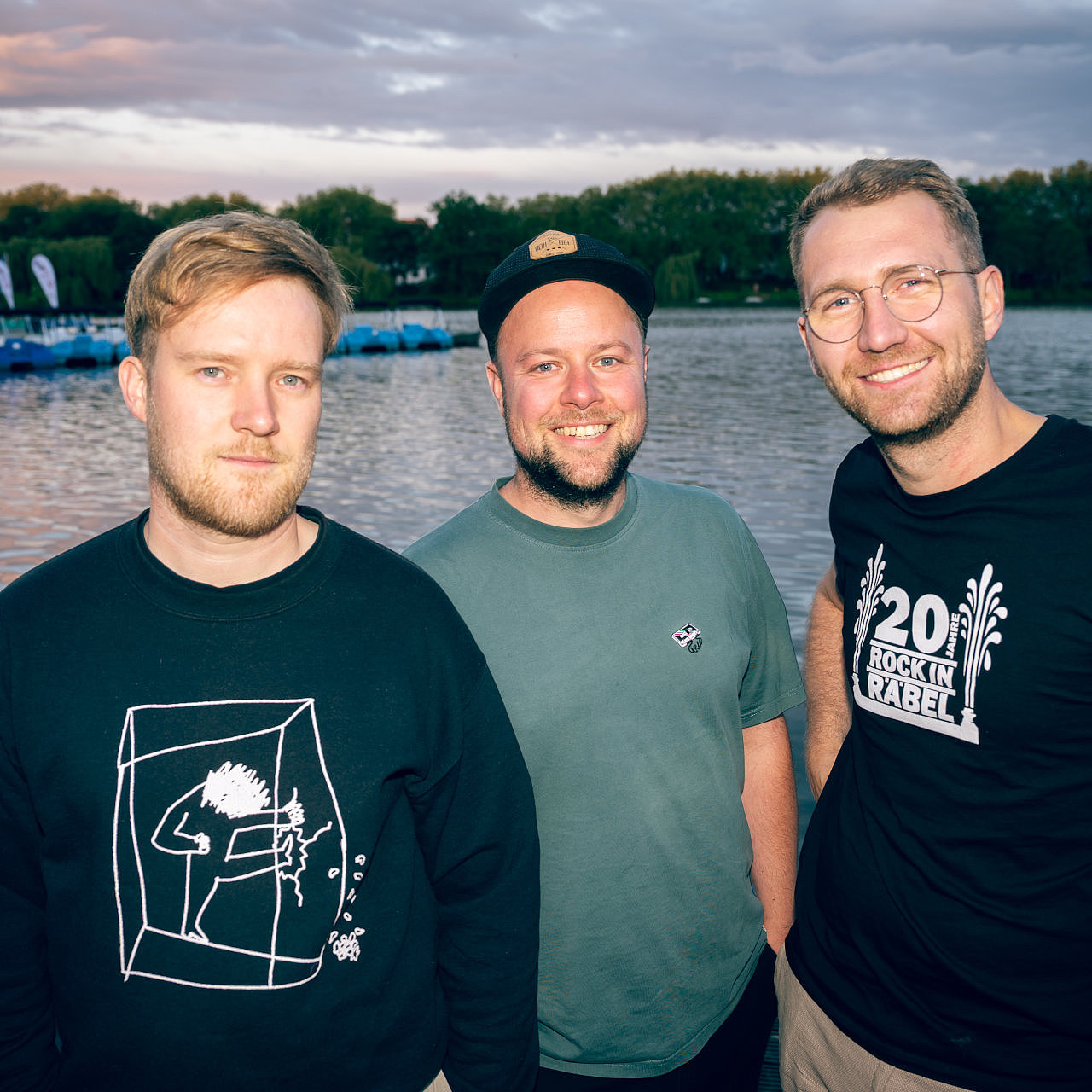 Die drei Bandmitglieder stehen vor einem See und blicken zur Kamera