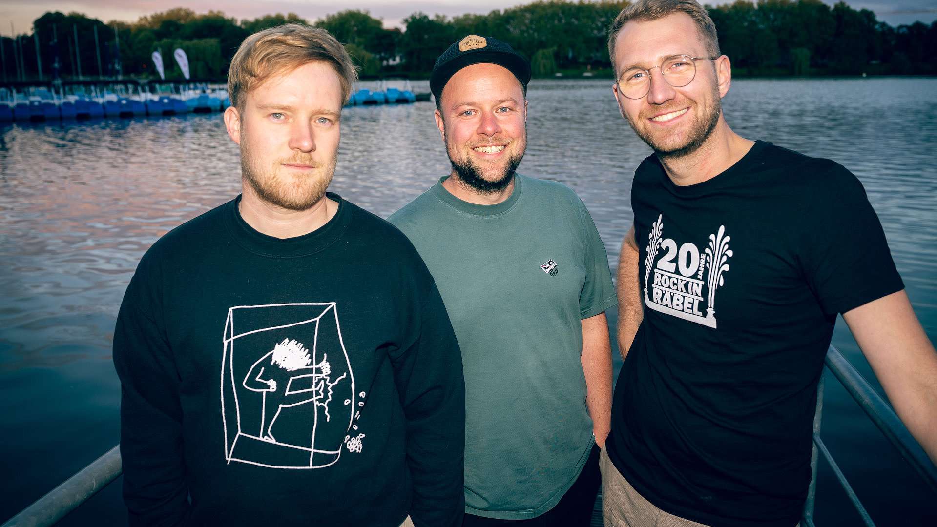 Die drei Bandmitglieder stehen vor einem See und blicken zur Kamera